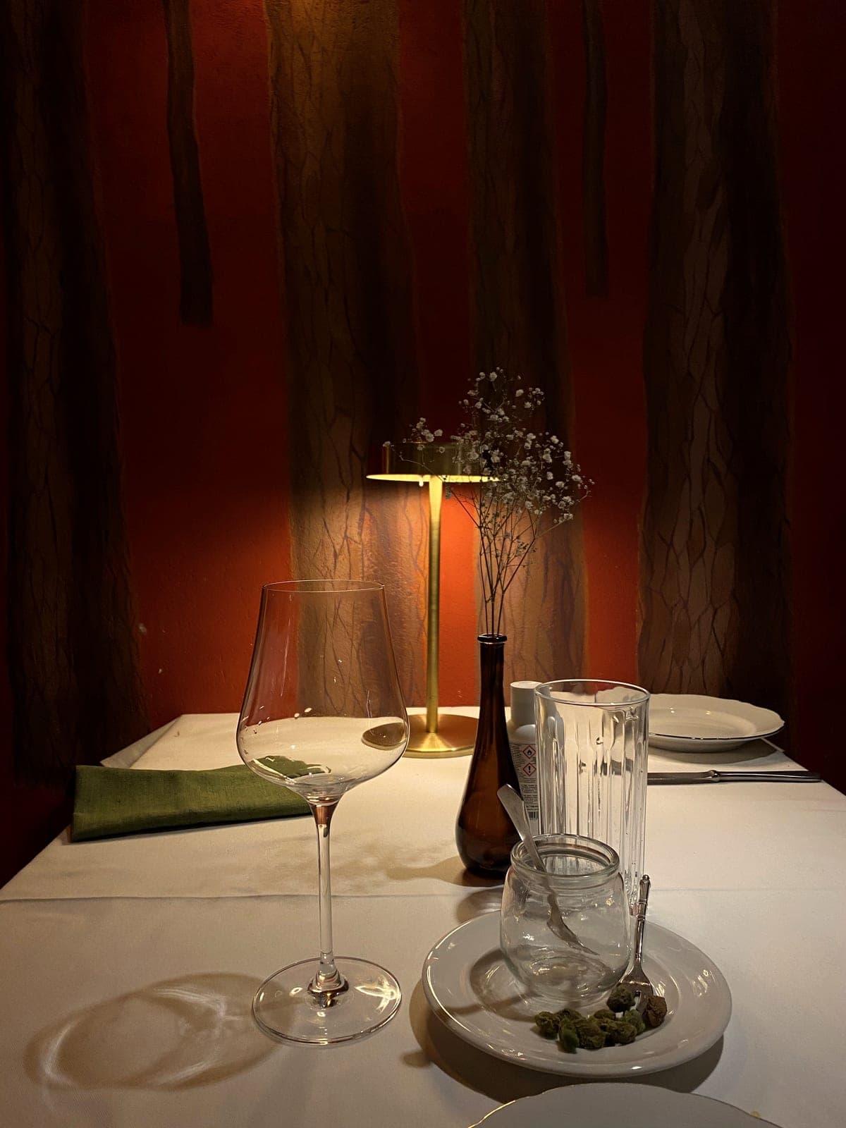A table laid for the evening: white linen, wine glasses and a small brass lamp against a deep red wall.